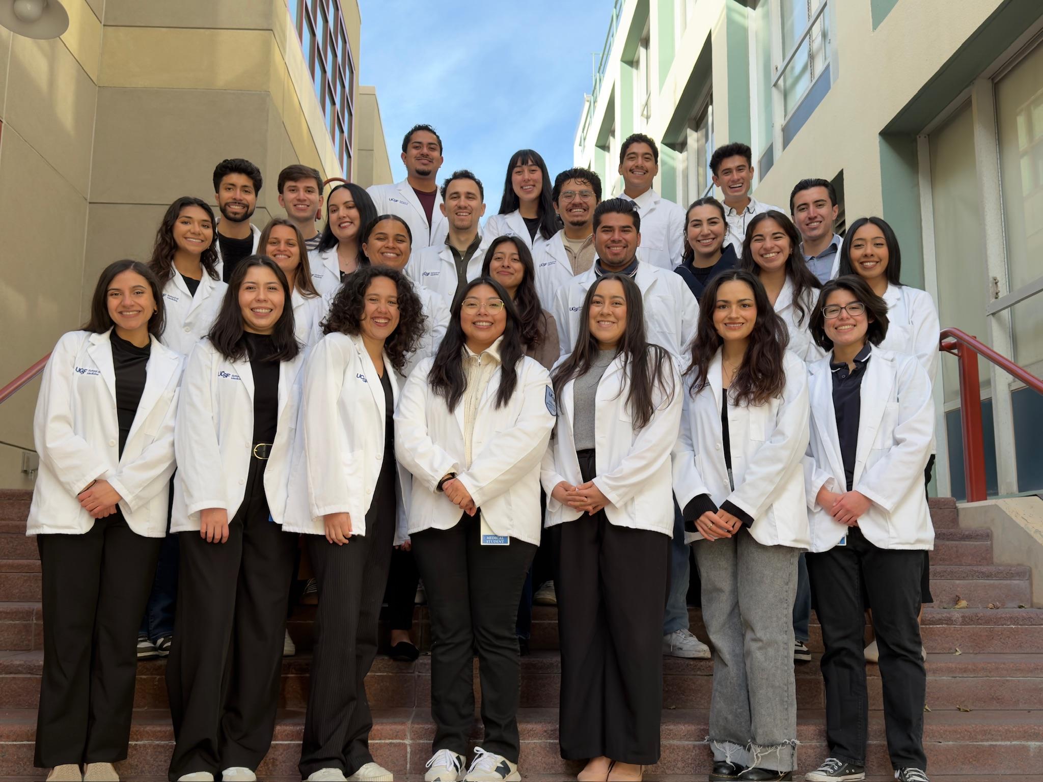 Group of UCSF medical students in white coats posing together on outdoor stairs.